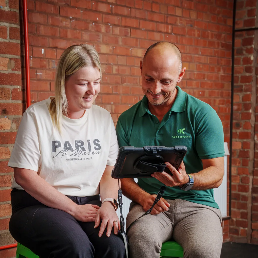 A man and woman sitting on green stools, reviewing information together on a tablet.