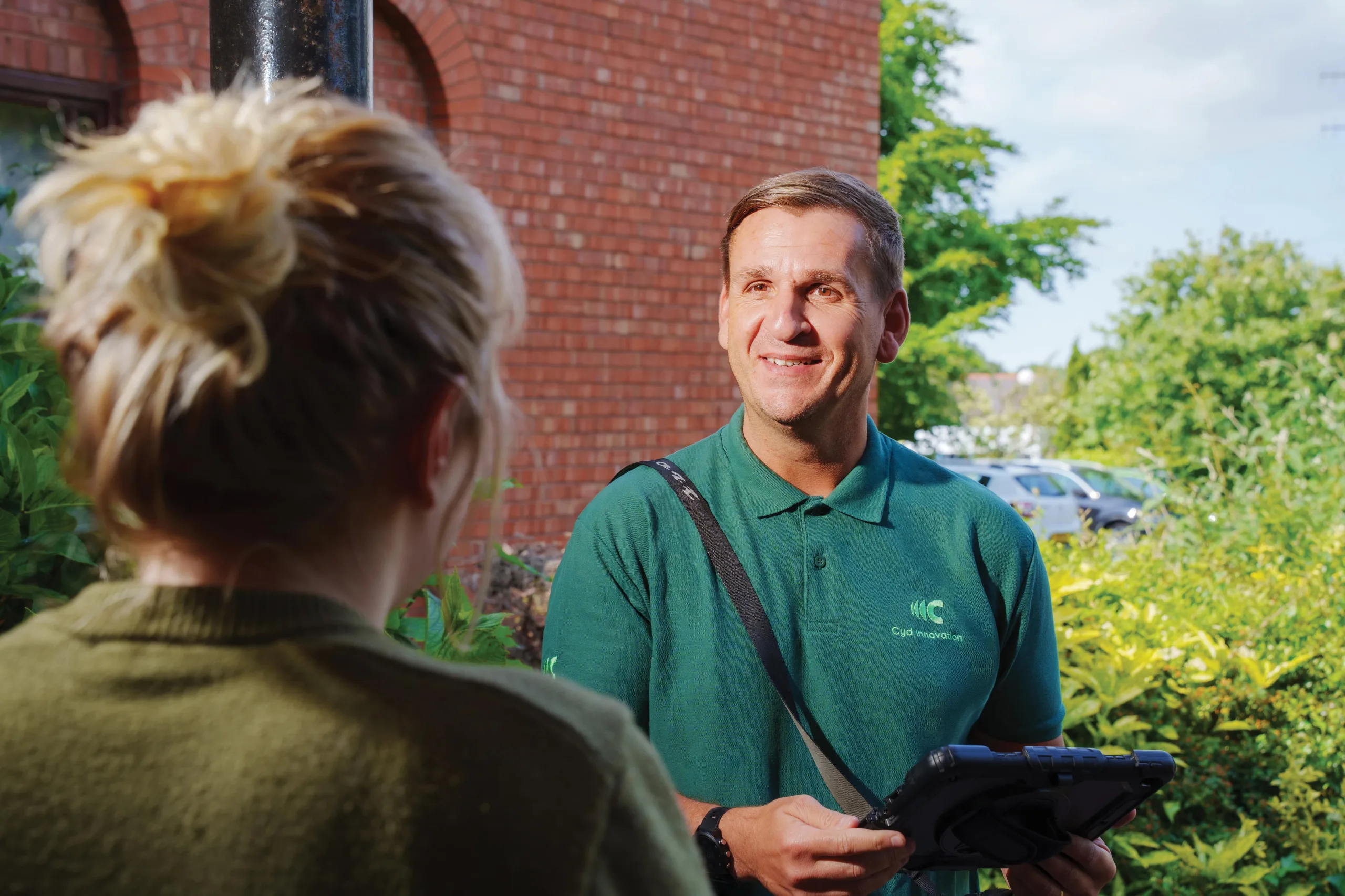 A man in a green shirt stands at the door, greeting a woman and holding a digital device.